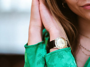 A close-up photograph of a woman with her hands up next to her head. She is wearing a green silk top and a watch is clearly visible on her wrist. Her hair is next to her hands.