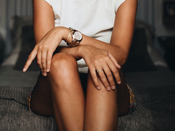 A close-up photo of a woman sat on a bed wearing a skirt. Her arms are relaxed and crossed above her legs, with a watch clearly visible on her left wrist.