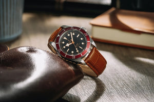 A close-up photo of a Tudor brand watch on a desk, with depth of field and other items scattered around including a book in the background. The watch strap is tan brown leather, and the watch face is black and the case bezel is red.