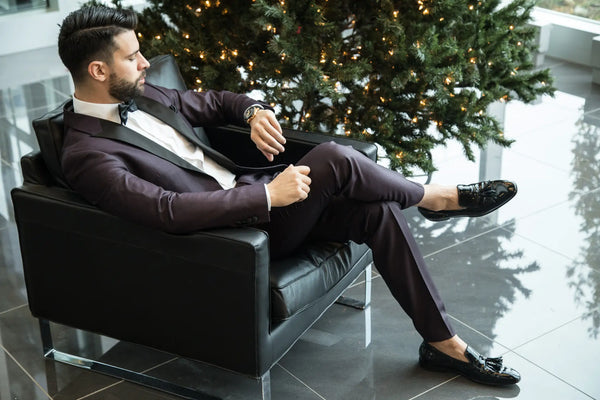 A photo of a man sat in a black leather chair wearing a dark purple tuxedo. He is looking at his watch on his left wrist and has his right leg crossed over his left.